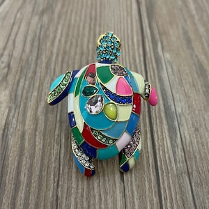 Puede incluir: Un broche de tortuga de mosaico colorido con un diseño de esmalte azul, verde, amarillo, rojo, rosa y blanco. La tortuga tiene un acabado dorado y está adornada con diamantes de imitación.