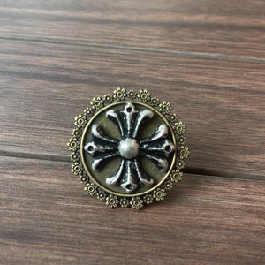 Può includere: Un ciondolo a forma di croce in argento e bronzo con un bordo floreale decorativo. La croce ha un design in rilievo ed è circondata da un cerchio di piccoli fiori.