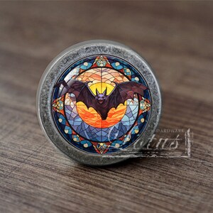May include: A silver cabinet knob with a stained glass design featuring a bat flying in front of a sunset. The stained glass design has a blue, orange, yellow, and red colour scheme.