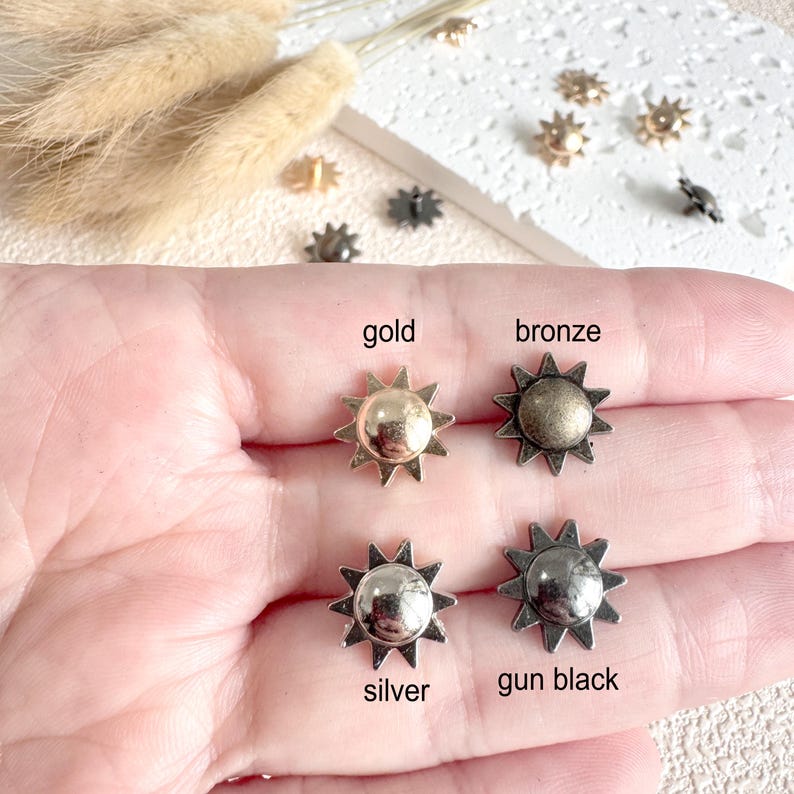 May include: Close-up of four sun-shaped metal studs in gold, bronze, silver, and gun black. The studs are held in a hand, with the colors labeled below each stud. Additional studs are scattered in the background.
