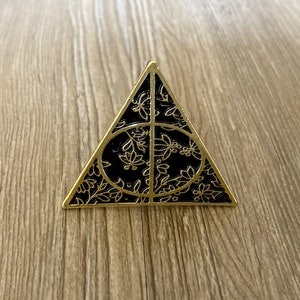 May include: A gold-colored metal pin in the shape of a triangle with a black interior. The black interior features a floral design within a circle and triangle, with gold-colored outlines. The pin is set against a wooden surface.