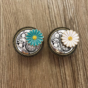 May include: Two silver and gold tone metal buttons with a flower design. The button on the left has a teal blue flower with a yellow centre. The button on the right has a white flower with a yellow centre.