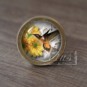 Könnte beinhalten: Ein runder, messingfarbener Schrankknopf mit einem Glaseinsatz, der einen Kolibri im Flug über gelben Blumen zeigt. Das Glas hat einen leichten, strukturierten, blasenartigen Effekt.