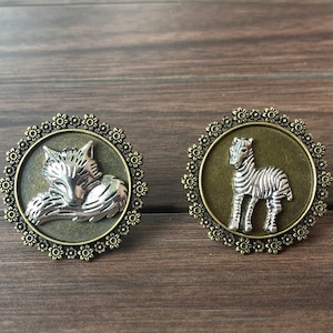 Könnte beinhalten: Zwei silberne und bronzene Broschen mit floralem Design. Eine Brosche zeigt einen schlafenden Wolf und die andere ein stehendes Zebra.