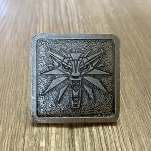 May include: A silver metal belt buckle with a wolf's head design. The wolf's head is surrounded by a stylized sunburst pattern.