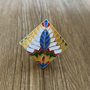 May include: A gold-coloured pin with a geometric design. The design features a blue, white and red stylised flower with green and yellow accents.