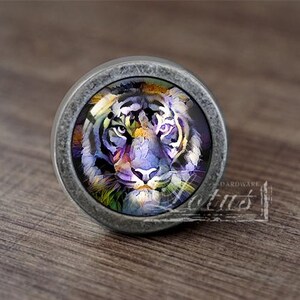 May include: A silver-toned cabinet knob featuring a colourful tiger face design. The tiger has purple, yellow and blue stripes.