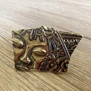 Pode incluir: Um ornamento de metal dourado com um rosto estilizado detalhado com os olhos fechados e desenhos florais. O ornamento tem uma forma irregular e uma superfície texturizada.