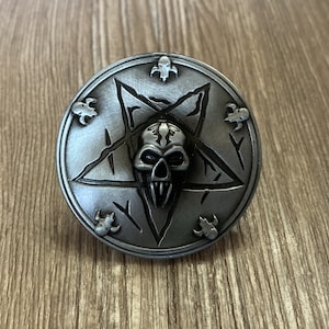 以下が含まれることがあります： 中央に五芒星と頭蓋骨のデザインが施された銀色の金属製のピン。頭蓋骨は鋭い歯と脅迫的な表情で詳細に描かれている。五芒星は5つの小さな頭蓋骨で囲まれている