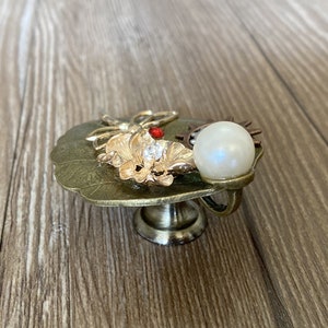 May include: A decorative gold-toned metal cabinet knob with a pearl, a butterfly, and a flower design. The knob has a round base and a stem.
