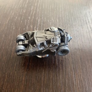 Puede incluir: Un modelo de coche de juguete de metal gris y detallado del Batmóvil de la franquicia Batman. El coche está en posición de accidente con las ruedas hacia arriba.