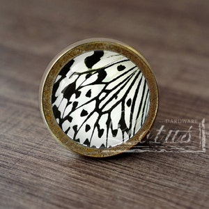 May include: A round, brass-colored cabinet knob with a black and white butterfly design. The knob has a rustic, aged look.