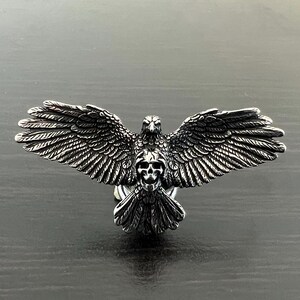 Peut inclure: Un aigle en argent aux ailes déployées, tenant un crâne dans ses serres. L'aigle est une sculpture tridimensionnelle détaillée.