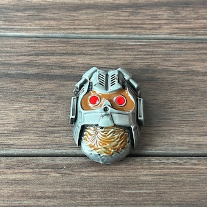 May include: A silver and brown metal pin featuring a character's face with red eyes. The character has a beard and a helmet with a detailed design.