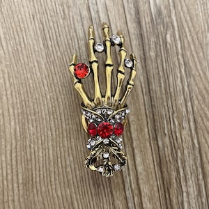 K&ouml;nnte beinhalten: Eine goldene Skeletthand-Brosche mit roten und klaren Strasssteinen. Die Hand h&auml;lt einen Strau&szlig; aus roten und klaren Strasssteinen.