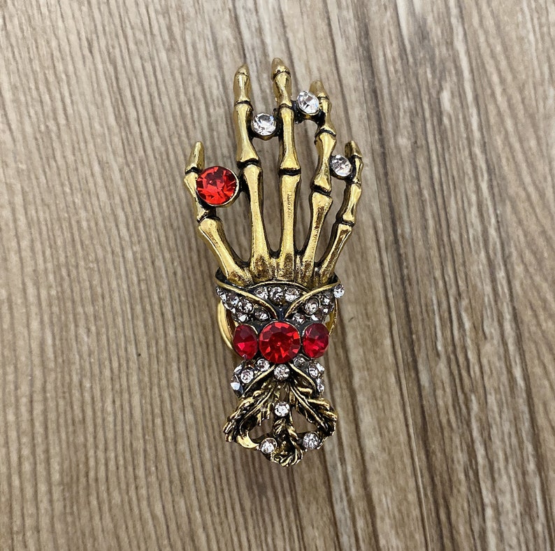 K&ouml;nnte beinhalten: Eine goldene Skeletthand-Brosche mit roten und klaren Strasssteinen. Die Hand h&auml;lt einen Strau&szlig; aus roten und klaren Strasssteinen.