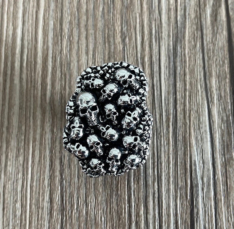 K&ouml;nnte beinhalten: Ein silberner Ring mit einem Design aus zahlreichen kleinen Totenk&ouml;pfen. Die Totenk&ouml;pfe sind fein detailliert und dicht gepackt. Der Ring hat einen gotischen oder Biker-Stil.
