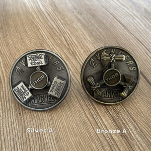 Peut inclure: Deux toupies en métal, l'une argentée et l'autre bronze, avec les mots "Avengers" et "Marvel" dessus. Les deux toupies présentent un motif de marteau de Thor.