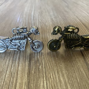 Pode incluir: Dois amuletos de motociclistas esqueleto, um prateado e outro dourado. As motocicletas têm pilotos esqueléticos detalhados e rodas.