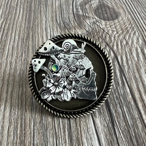 May include: A silver skull brooch with a detailed design of mushrooms, flowers, and a snail. The brooch has a dark brown background and a rope-like border.