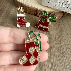 May include: Three gold-tone Christmas-themed brooches. One is a red stocking with white trim and gold and white rhinestones. Another is a red stocking with a green bow and gold and white rhinestones. The last is a red bell with green holly and gold and white rhinestones.