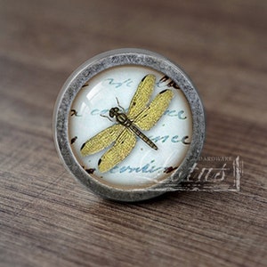 May include: A round, silver-toned cabinet knob with a glass insert featuring a yellow dragonfly on a white background with blue and gray script.