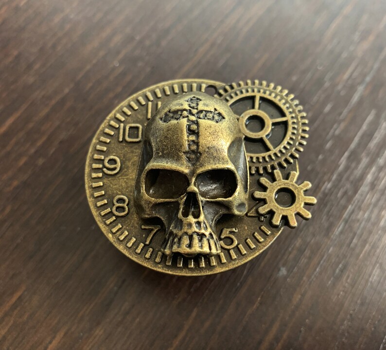 May include: A gold-toned skull pendant with a cross design on the forehead, surrounded by a circular clock face with Roman numerals. The pendant is attached to a gear-shaped charm.