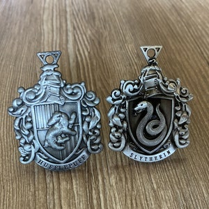 May include: Two silver metal Harry Potter house crests. The crest on the left is for Hufflepuff and features a badger. The crest on the right is for Slytherin and features a snake.