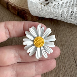 May include: A white daisy flower brooch with a yellow centre. The brooch has white petals and a yellow centre. The daisy is held in a hand. The brooch is a floral accessory.