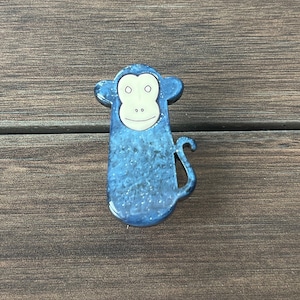 Puede incluir: Una figurita de mono de cerámica azul con una cara blanca y una sonrisa simple. El mono tiene una cola larga y está de pie sobre una superficie de madera.