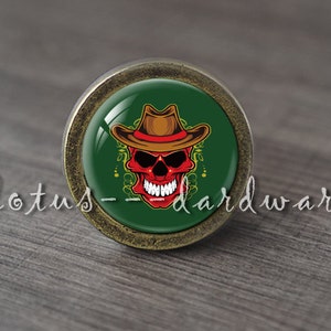 Puede incluir: Un pomo de armario redondo de color dorado antiguo con un inserto de vidrio verde. El inserto de vidrio presenta una calavera roja con un sombrero de vaquero marrón.