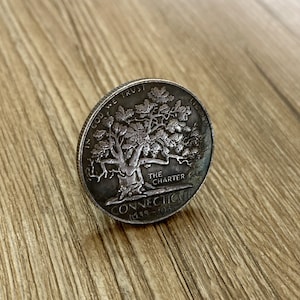Op de afbeelding: Een zilveren munt met de woorden "In God We Trust" en "The Charter Oak". De munt heeft ook de woorden "Connecticut 1639 - 1989" erop.