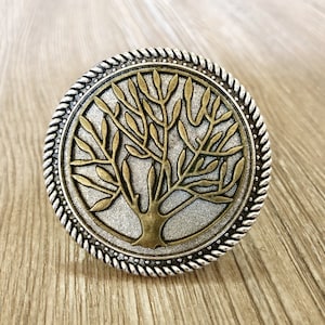 以下が含まれることがあります： 生命の木のデザインが施された、銀と金の丸い金属製のノブ。ノブはロープのような縁取りと質感のある表面をしています。