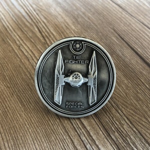 Peut inclure: Une pièce de monnaie en métal argentée avec un design de vaisseau spatial TIE fighter. La pièce porte les inscriptions "TIE FIGHTER" et "SPECIAL FORCES".