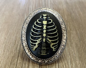 May include: A silver-toned brooch with a black cameo featuring a white ribcage design.