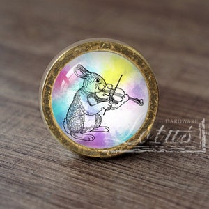 May include: A round, gold-colored cabinet knob with a colorful watercolor background and a black and white illustration of a rabbit playing the violin.
