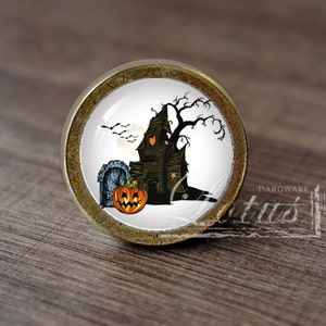 Pode incluir: Um botão de armário redondo de ouro antigo com um inserto de cerâmica branca que apresenta uma cena assustadora de Halloween. A cena inclui uma casa assombrada, uma abóbora talhada, uma lápide e uma árvore nua com corvos voando por cima.
