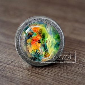 Può includere: Un pomello per armadio rotondo di colore argento con un inserto di vetro che presenta un dipinto colorato di due volti di lupo.