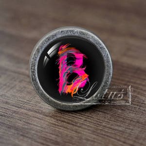 May include: A round, silver-coloured cabinet knob with a black background and a pink, orange, and blue letter "B" painted on it.