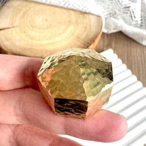 May include: A faceted, golden-colored object with a hammered texture is held in a hand. The object has a geometric shape and reflects light. A wooden slice and white ribbed surface are in the background.