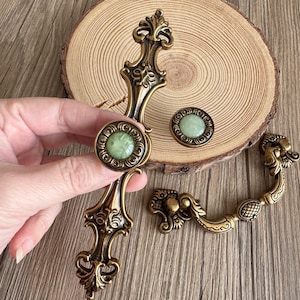以下が含まれることがあります： アンティーク真鍮製のキャビネット金具セット。装飾的な引き手とノブが含まれています。引き手は、中央に緑色の石が付いた長い装飾的なデザインです。ノブにも緑色の石が中央にあります。セットには、2つの引き手と2つのノブが含まれています。