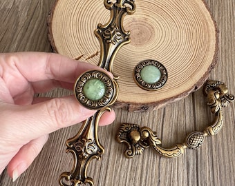 May include: A collection of antique-style brass cabinet hardware featuring ornate scrollwork and central circular inlays of pale green stone. The set includes a long vertical handle, a matching round knob, and a curved drop-style drawer pull.