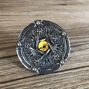 May include: A silver ring featuring a pentagram design with a yellow eye at its centre. The ring is intricately carved and circular in shape.