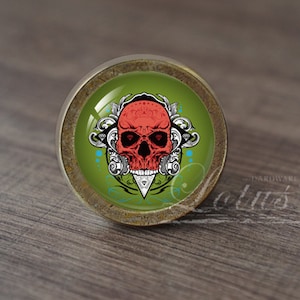 Op de afbeelding: Een ronde, antieke messing kastknop met een groene glazen inzet met een rode schedelgrafiek met witte details en een zwarte achtergrond. De schedel is omgeven door een zilveren bloemendesign.