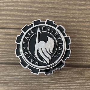 May include: A black and silver metal pin with a gear-like design. The pin features the words "Fairy Tail" written in a circle around a stylized wing with a flame design.