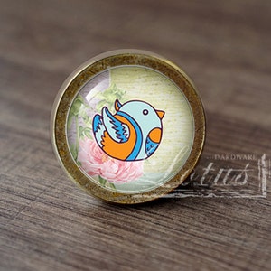 Puede incluir: Un pomo de armario redondo de color dorado con un inserto de vidrio que presenta un pájaro de dibujos animados azul y naranja sobre un fondo floral.