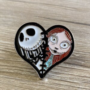 May include: A black and white heart-shaped pin with a black and white image of Jack Skellington from The Nightmare Before Christmas on the left side and a red-haired Sally on the right side. The pin is broken in the middle and stitched together.