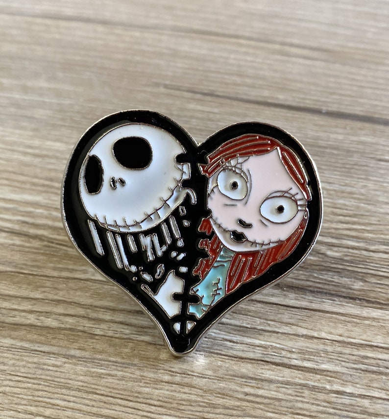 May include: A black and white heart-shaped pin with a black and white image of Jack Skellington from The Nightmare Before Christmas on the left side and a red-haired Sally on the right side. The pin is broken in the middle and stitched together.