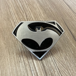 Op de afbeelding: Een zilveren gesp met een zwart Batman-logo in een Superman-symbool.