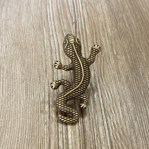 Peut inclure: Une figurine de lézard en métal doré avec un design texturé et écailleux. Le lézard est posé sur ses pattes tendues et sa queue enroulée.